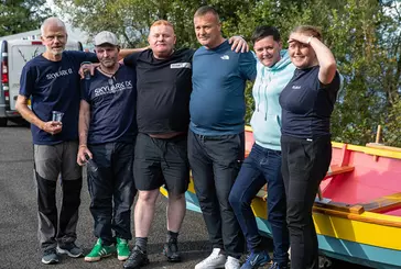 A group of people wearing Skylark IX t-shirts pose together in front of a brightly painted wooden rowing boat