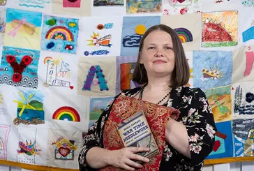 A woman stands in front of a patchwork quilt inspired by the Skylark IX and memories of lockdown