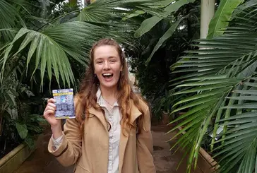 A young woman with long blonde hair standing in the Palm House at Kew Gardens, holding a scratchcard