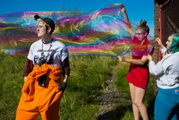 Three young people stand outside waving a rainbow coloured piece of fabric