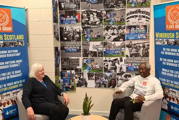 Two older people talking. Behind them are banners for the Windrush Scotland project