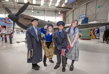 Four children, dressed in WW2 attire and holding union flags, stand in front of a Spitfire at the RAF Museum Midlands.