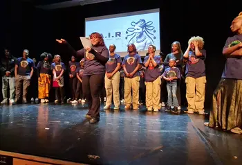A large group on stage with a person reading from a sheet of paper in the foreground