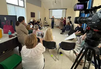 People sit on chairs in a circle watching someone give a presentation. In the foreground a video camera films them.