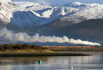 A steam train travelling next to water with snowy mountains behind
