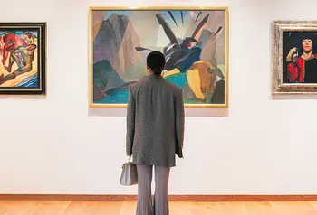 A young person looking at one of three paintings on a gallery wall. The person has their back to us.