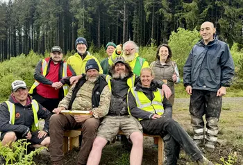 A group of men and women in high vis jackets and outdoor attire gather around a newly built bench on a wooded hillside.