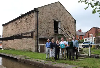 A group of smiling people, holding a blue Heritage Fund roundel, stand between the canal and the historic wharf building behind them.