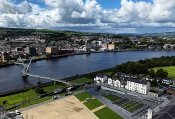 A public square with buildings next to it on the edge of a wide river, the city on the other side of the river is connected by a large white bridge