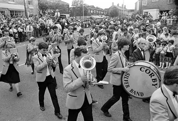 A black and white image of a brass band marching through the streets of a packed town centre.