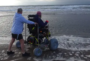 Man pushing person using large wheelchair next to river