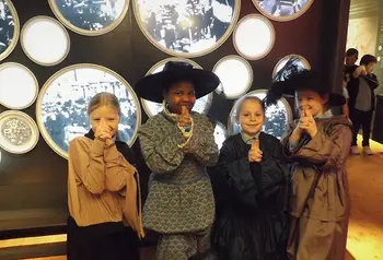 Four children face camera smiling. They are dressed in historic outfits. They are near an exhibition. 