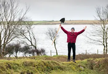 Person yn sefyll ym myd natur yn edrych yn hapus wrth ddal rhaw yn yr awyr
