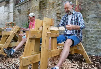 Two people sitting outside carving wood at woodworking benches