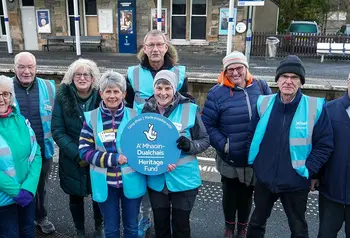 A group of people wearing cold weather gear stand on the platform of a rural train station holding a sign 