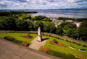 Bo'ness war memorial is at the centre of the frame and behind it there are trees and houses. In the distance is the Firth of Forth.