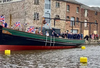 A crowd of people, some in maritime uniforms, gathered around a ship's hull docked next to harbour buildings. The ship is flying Union Jack flags and nautical bunting.