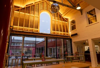 A room with a high vaulted ceiling, a large glitter ball and trestle style tables. The room is warmly lit. Large glass doors look out onto a courtyard. 