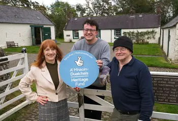 Three people standing at a gate. Behind them are white farm buildings. They are holding a blues circular The NAtional Lottery Heritage FUnd sign