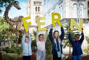 four children hold up large letters spelling FERN, the name of the diplodocus skeleton on display behind them