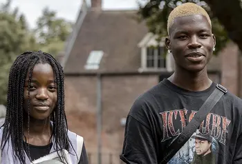Two young black people wearing casual clothes stand outside with a historic brick building in the background
