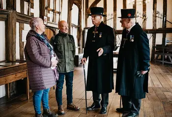 two visitors are given a tour of a timber framed hall by two older gentlemen wearing long black coats at traditional hats