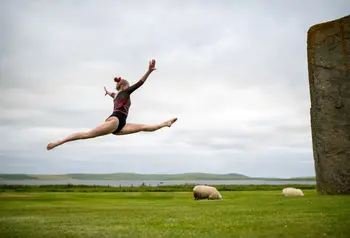 A gymnast leaps through the air in a field on Orkney, with sheep and standing stones nearby.