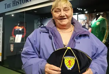 A woman in a purple jacket holding a Scotland international football cap with the name Janie Houghton embroidered on the peak