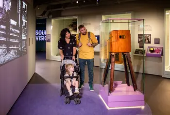 A man and a woman push a child in a buggy as they take a closer look at a pioneering film camera from the early days of the technology. A black and white film of the era is projected on the wall to their left.