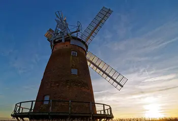 Looking up at a red brick windmill with large white sails, as the sun sets to the right.