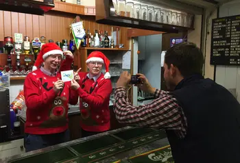 A person taking a photo of two people behind a bar wearing red Christmas hats and jumpers.