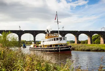 A boat on a waterway with a bridge in the background and plants visible on the banks of the water