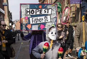 a parade in a town high street, one person holds up a sign with 'hope street' on it and a young person in a homemade mask shakes maracas