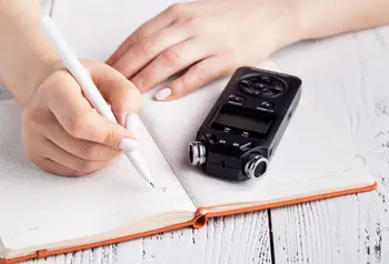 A voice recorder on a table next a a notebook with someone getting ready to take notes