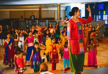 A woman on a stage leading an Indian dance class for children, everyone is wearing colourful traditional dress