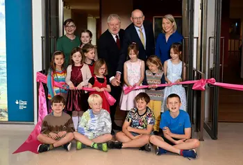 A group of children and adults sit and stand as they pose for a photo while cutting a ribbon.
