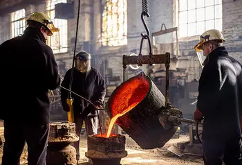 Three people stand inside a factory casting a large bell. Molten metal pours from a container held in the air by a hoist