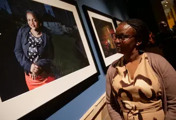 On the left is a framed photograph of a woman on the right there is a woman in a light brown dress looking at the picture