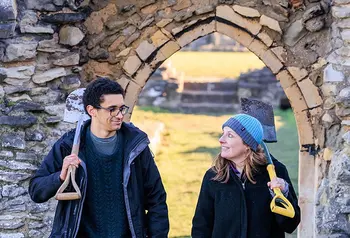Two people holding shovels and dressed for cold weather walk through a doorway in a wall