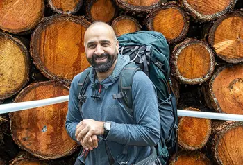 A man from a Pakistani family background on a outdoor walk, wearing hiking gear. He stands in front of a pile of logs