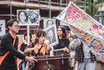 Two people of Chinese heritage play a drum in a procession, while people in the background hold placards with photos of Chinese relatives