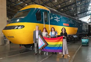 Four people stand in front of a train engine and hold a progress pride flag