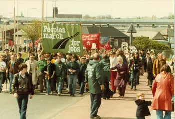 A group of people on a march through an industrial areas. They are holding a large banners, one reads 'People's March for Jobs' 