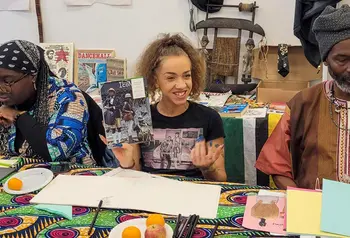 Three black people take part in a workshop on black history. All three people have dark skin of various shades, the focus of the image is a woman holding a magazine with black people on the cover