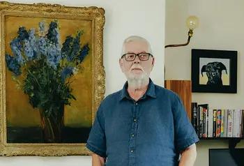 Richard Kennedy stands in a living room in front of paintings and a bookshelf