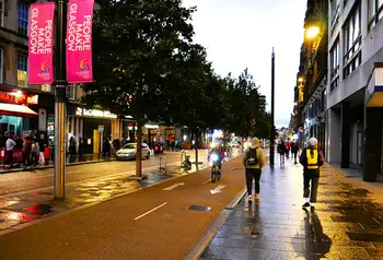 The western end of Sauchiehall Street at dusk: on the right people are walking and cycling by a People Make Glasgow banner and on the left a group of people are standing outside a bar