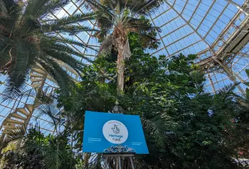 Palm trees and plants inside a glassed roofed building. A board with the Heritage Fund logo is on display on an easel. 