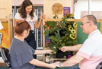 Two people sit in chairs holding hands with their other hands touching the leaves of a potted plant. Wires run from the plant to pads on the people's wrists. A smiling person in a Sense t-shirt watches.