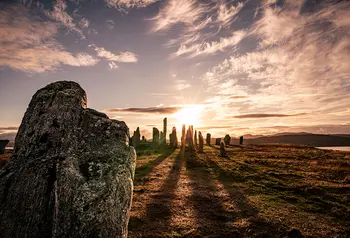 In the foreground to the left is a standing stone long shadows reach out to it from a group of standing stones behind. The sun is setting with orange tinged light filtering through the cluster of stones