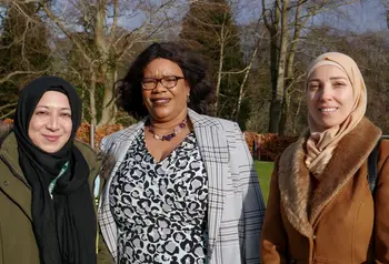 Three women from diverse ethnic communities are stood and smiling at the camera